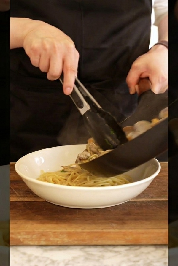 Un cuisinier en tablier noir utilisant une pince pour servir les pâtes aux palourdes dans un bol blanc.