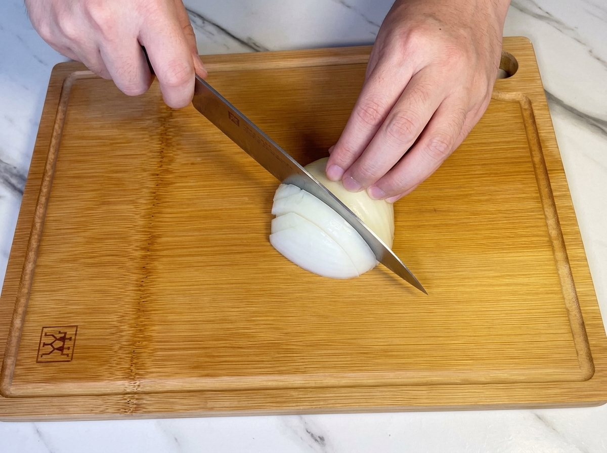 Mains utilisant un couteau de chef pour émincer un demi-oignon blanc sur une planche en bois.