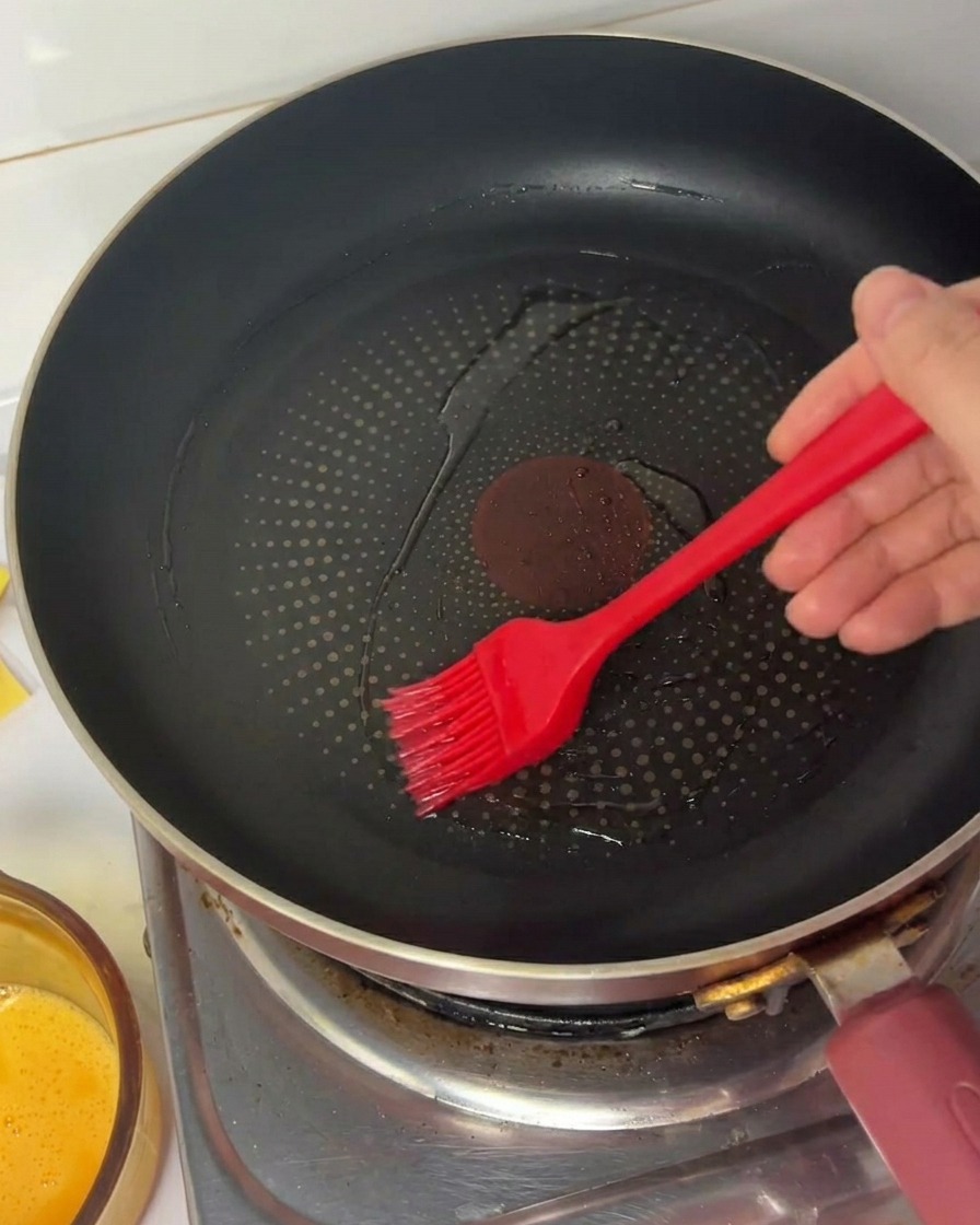 Une main utilisant un pinceau en silicone pour étaler lhuile dans une poêle antiadhésive noire.