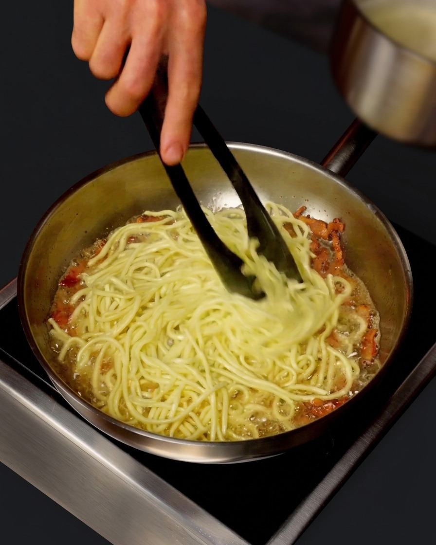 Une petite casserole versant de leau de cuisson chaude dans une poêle remplie de spaghettis mélangés avec des pinces.