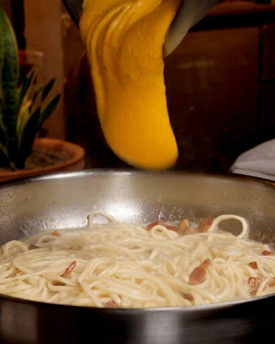 Un mélange épais dun orange vif à base dœufs et de fromage versé depuis un bol en métal sur une poêle de spaghettis.