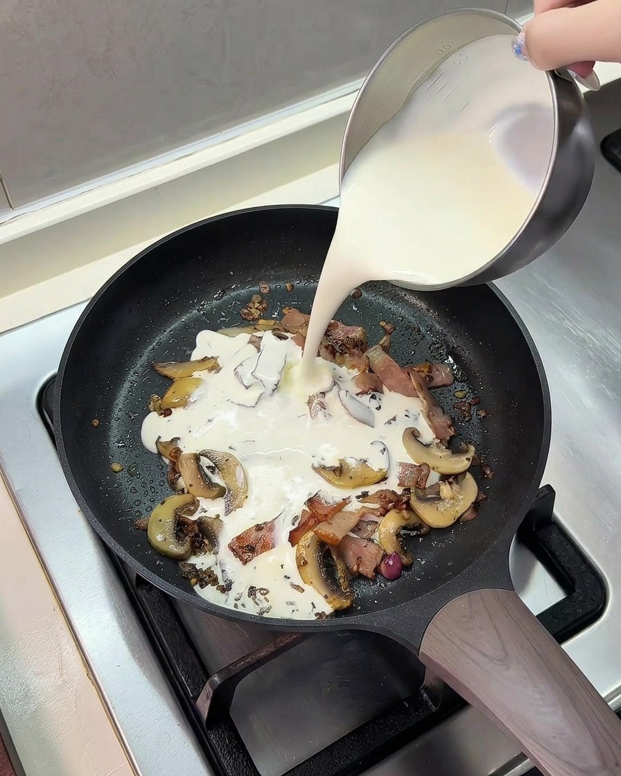 Un mélange de lait et de crème entière versé depuis un pichet en métal dans la poêle garnie de champignons et lardons.