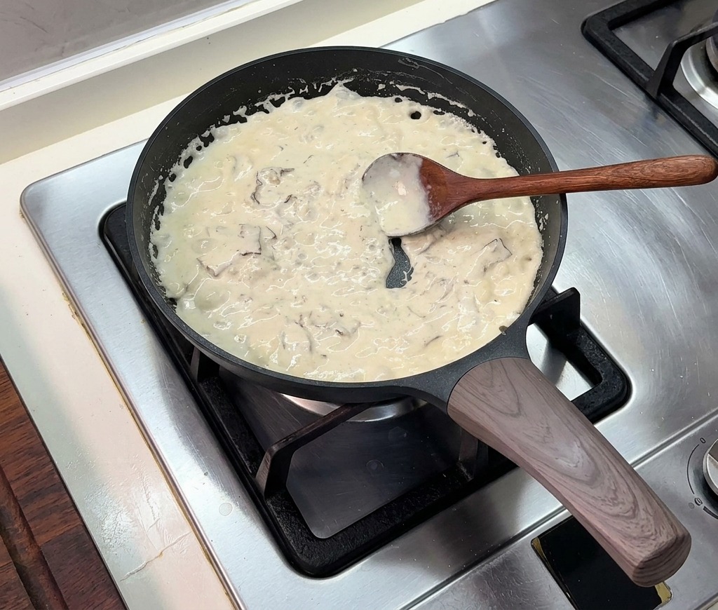 Une sauce à la crème épaisse mijotant dans une poêle noire, remuée à la cuillère en bois.