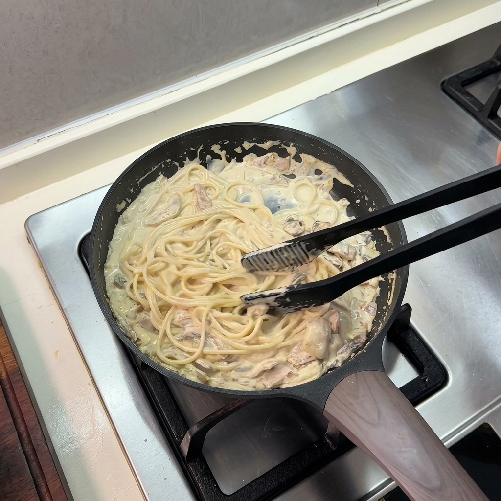 Des pinces de cuisine noires mélangeant les spaghettis cuits à la sauce crémeuse dans la poêle.