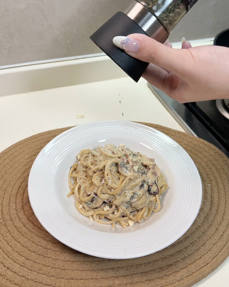 Une main râpant du fromage et saupoudrant des herbes sur une belle assiette de pâtes fumantes aux champignons et lardons.