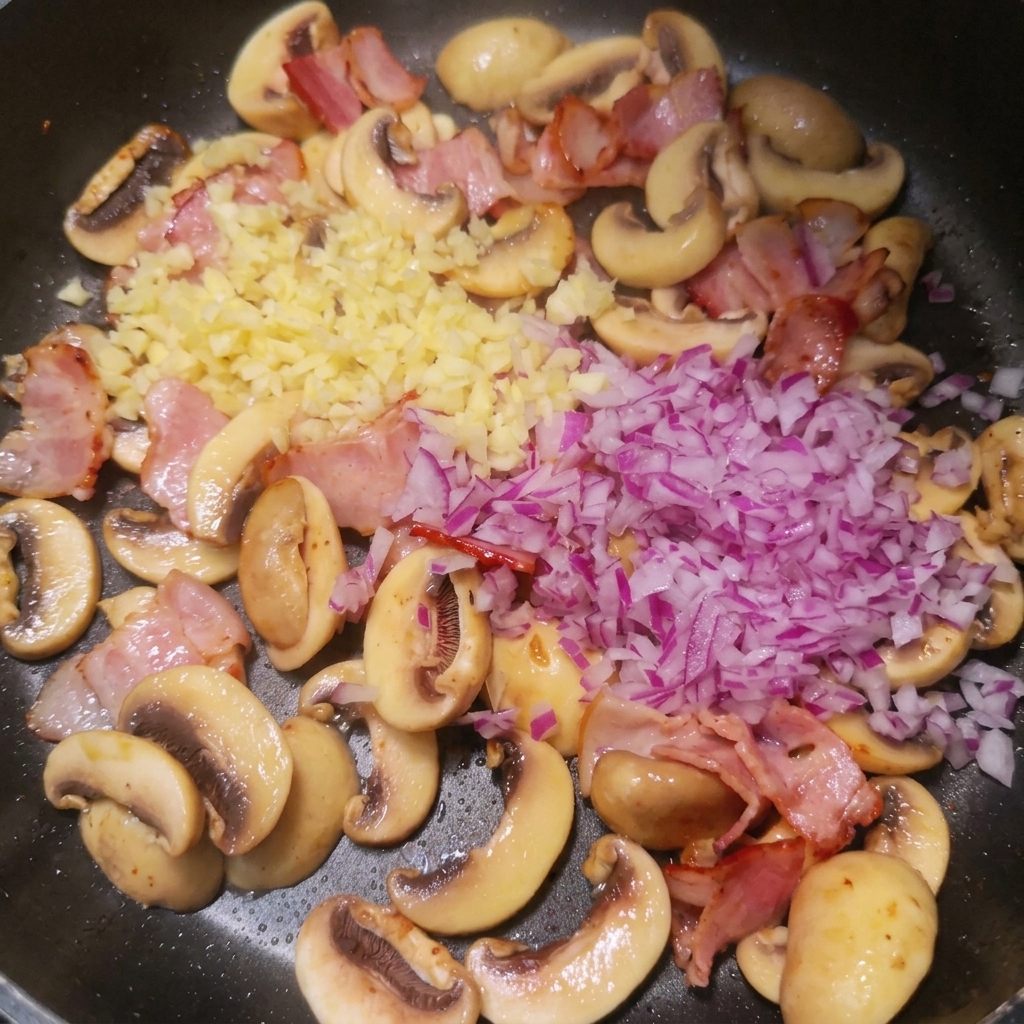Ail et oignon rouge hachés déposés sur le mélange bacon et champignons dans la poêle.