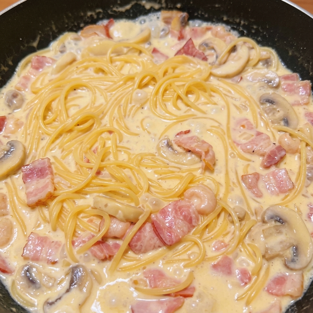 Spaghetti cuits mélangés à la sauce crémeuse au bacon et champignons dans une sauteuse.