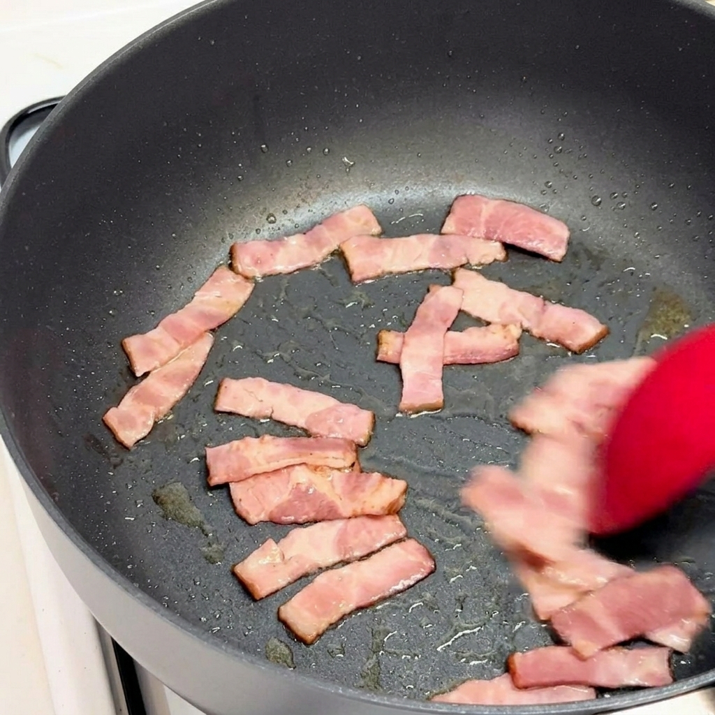 Morceaux de bacon sautés dans une poêle noire avec une spatule rouge.