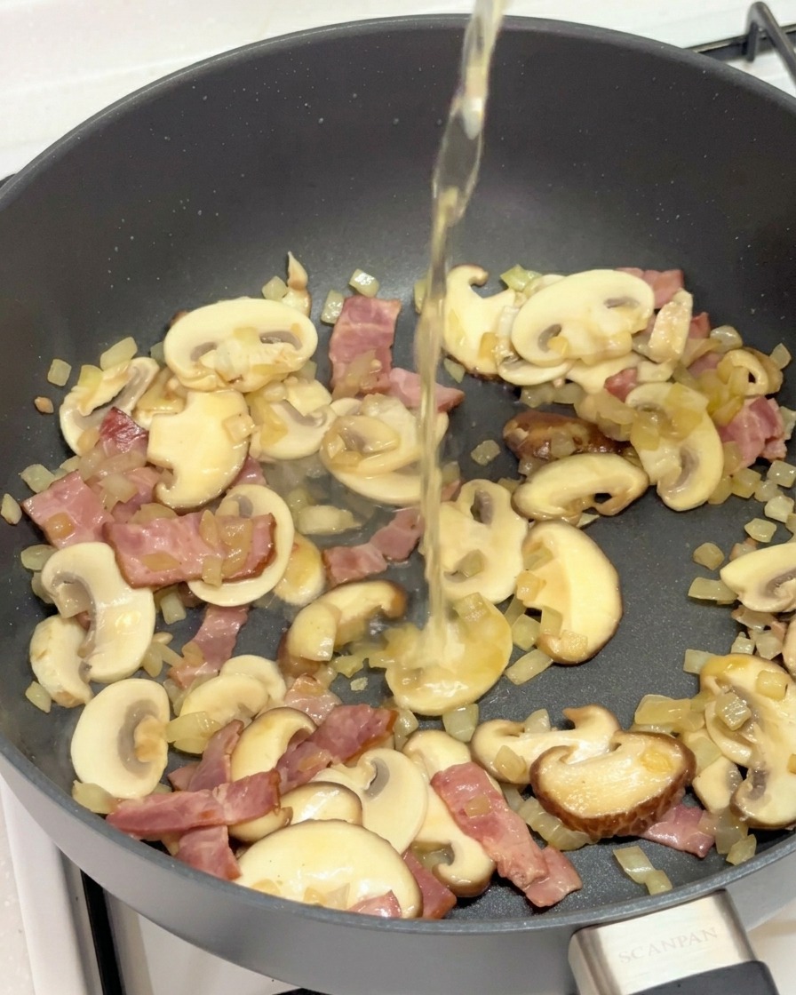Liquide clair versé dans une poêle remplie de champignons, bacon et oignons.