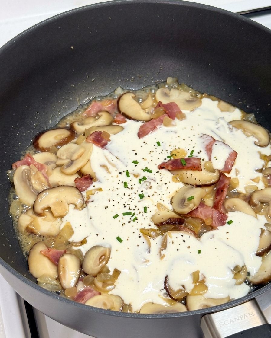 Crème mijotant dans une poêle avec champignons et bacon, parsemée de poivre.