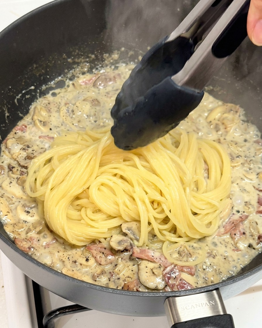 Pince déposant des spaghettis dans une poêle remplie de sauce à la truffe.
