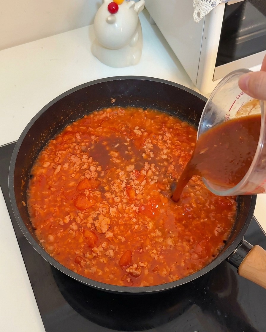 Un liquide sombre versé dun verre doseur dans une poêle de viande et de tomates mijotées.