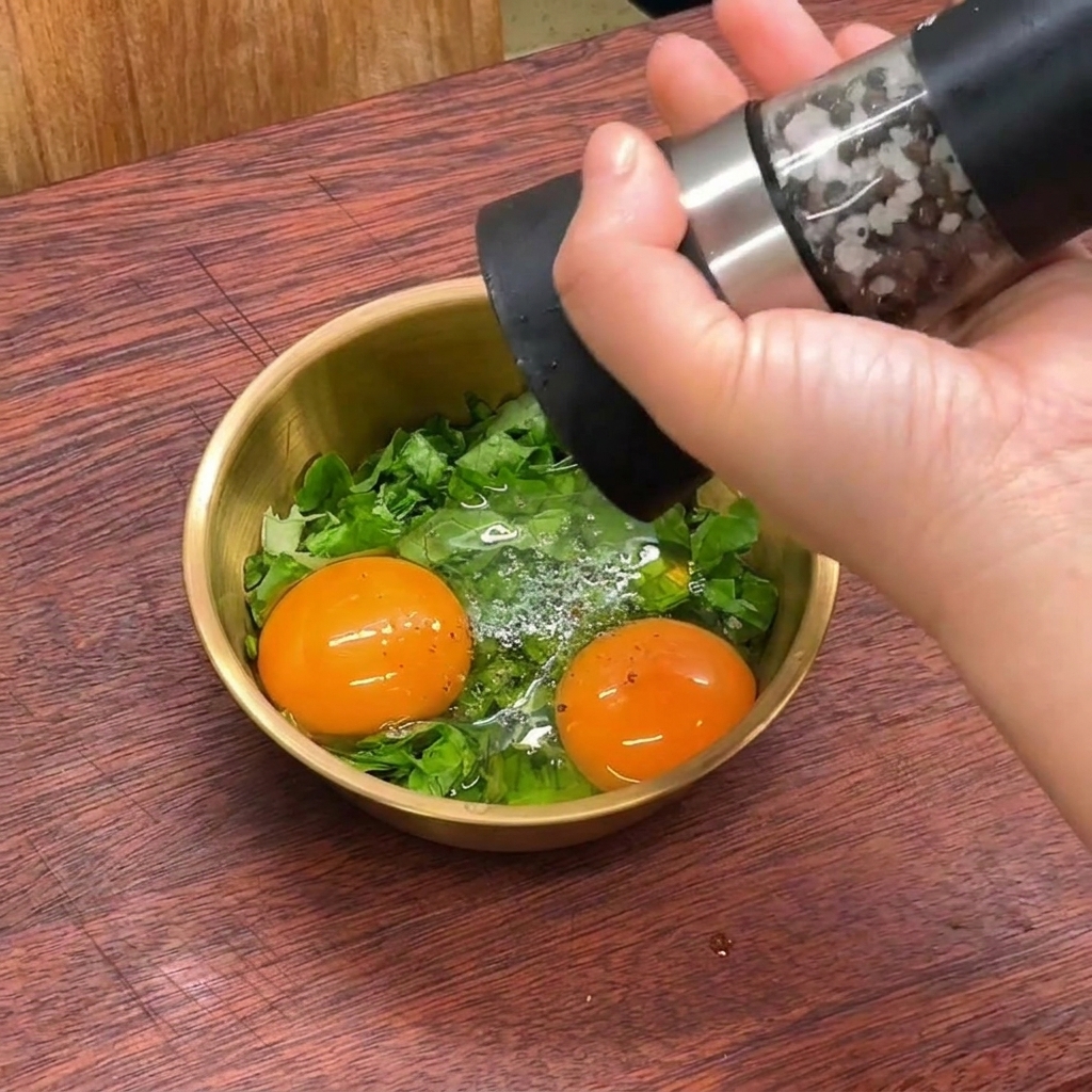 Une main utilisant un moulin à poivre pour moudre du poivre noir frais dans un bol contenant des œufs crus et de la laitue verte émincée.