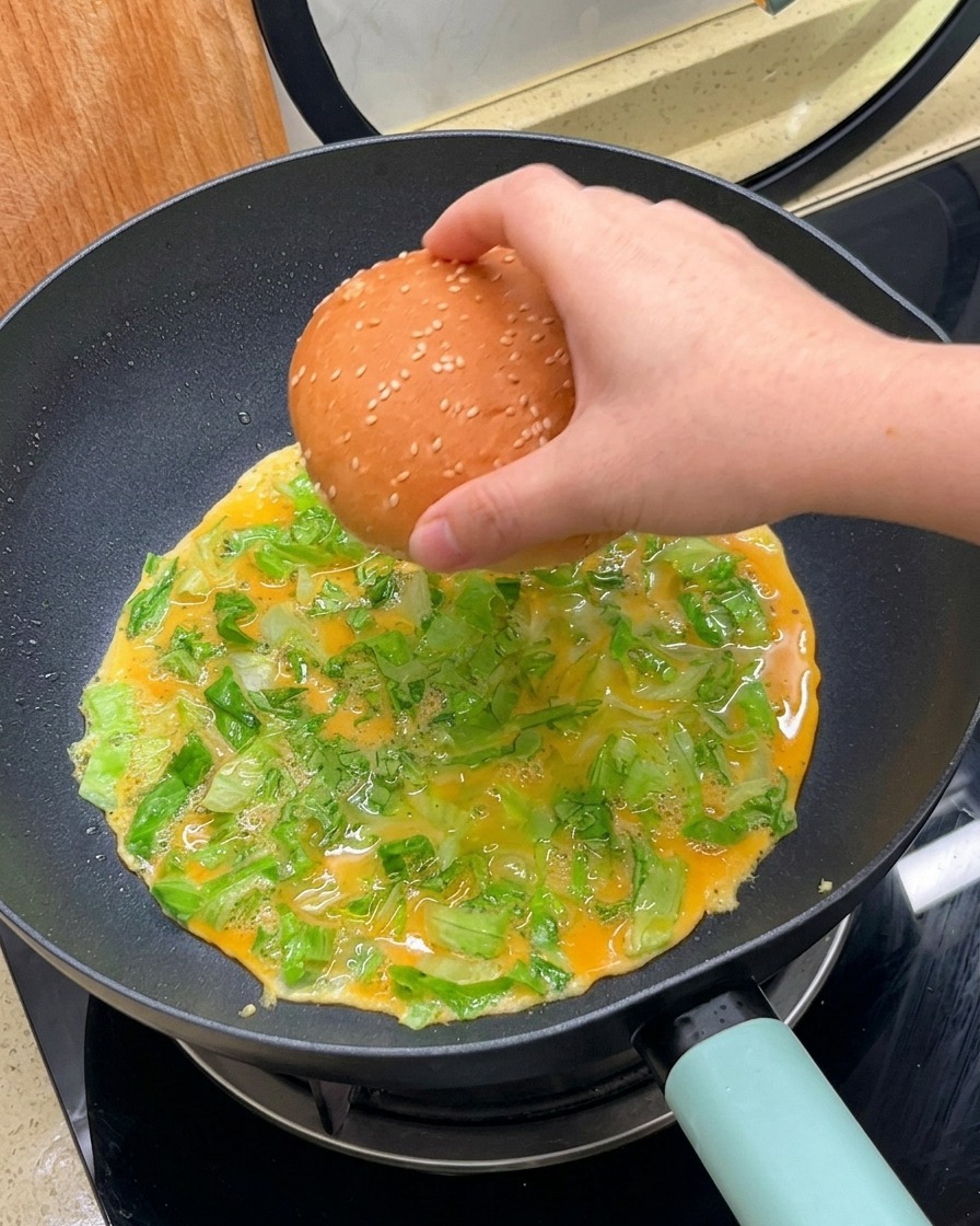 Une main plaçant un pain burger au sésame face contre lomelette en cours de cuisson dans la poêle.