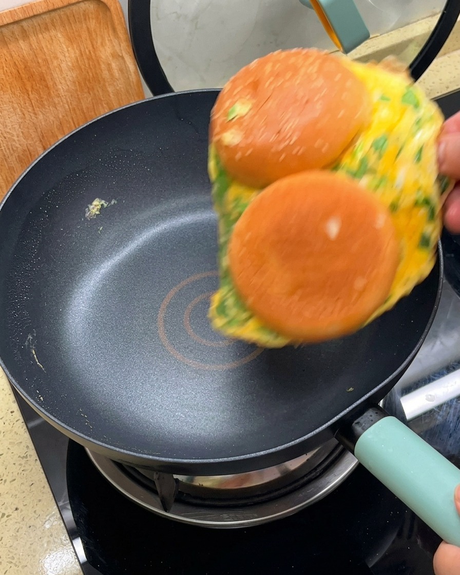Une omelette avec deux moitiés de pain burger fixées dessus, retournée dans une poêle antiadhésive.