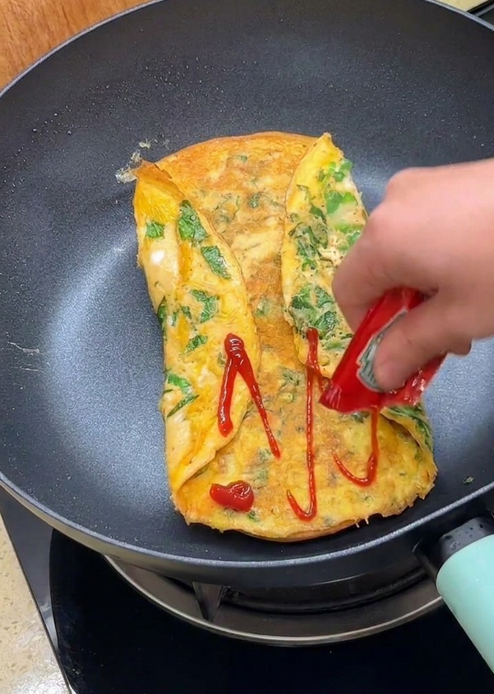Une main versant du ketchup sur la surface de lomelette pliée dans la poêle.