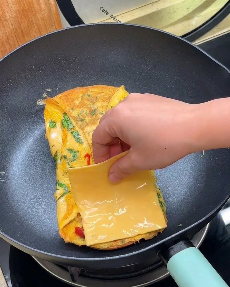 Une main plaçant une tranche de fromage jaune sur lœuf plié dans la poêle.