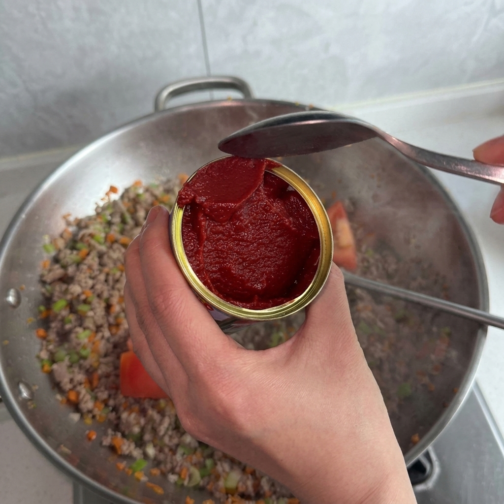 Une personne tenant une boîte de concentré de tomates et utilisant une cuillère pour lajouter au mélange viande et légumes.