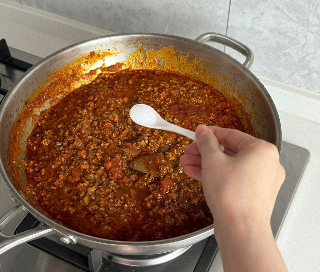 Une main ajoutant du sel blanc avec une petite cuillère dans une sauce rouge frémissante.