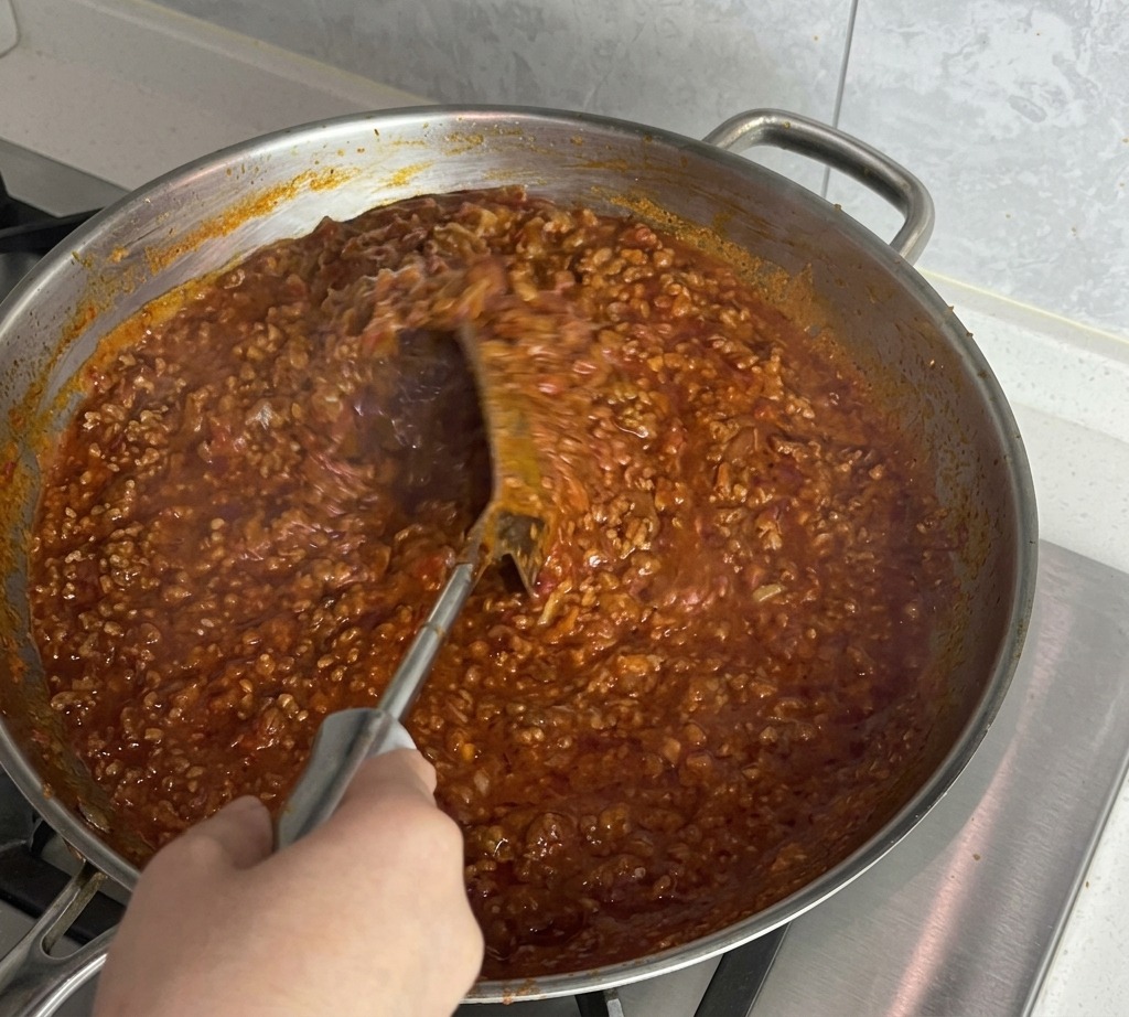 Une main utilisant une spatule pour mélanger vigoureusement une sauce bolognaise épaisse.