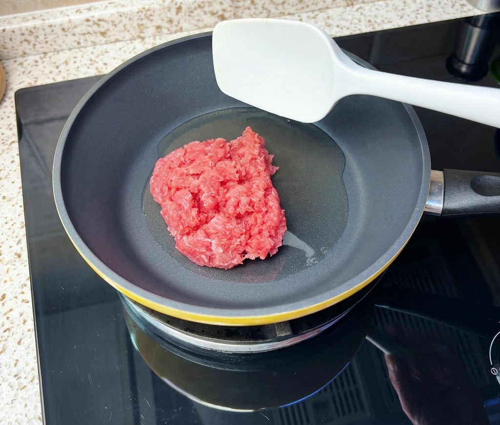 Un bloc de bœuf haché cru déposé dans une poêle chaude avec de lhuile à laide dune spatule.