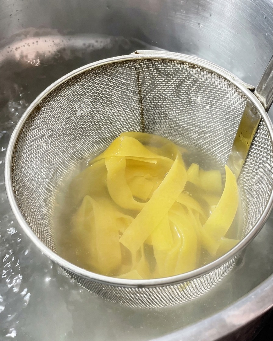 Larges rubans de pâtes cuisant dans une passoire métallique plongée dans une casserole deau bouillante.