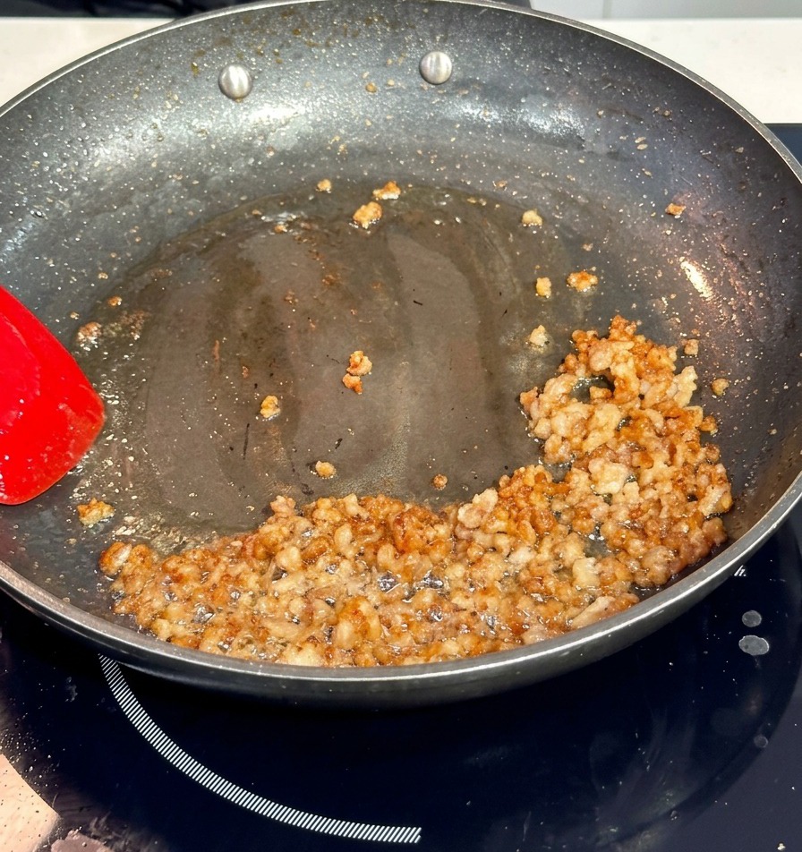 Viande de porc hachée dorant dans une poêle antiadhésive sombre, remuée avec une spatule rouge vif.