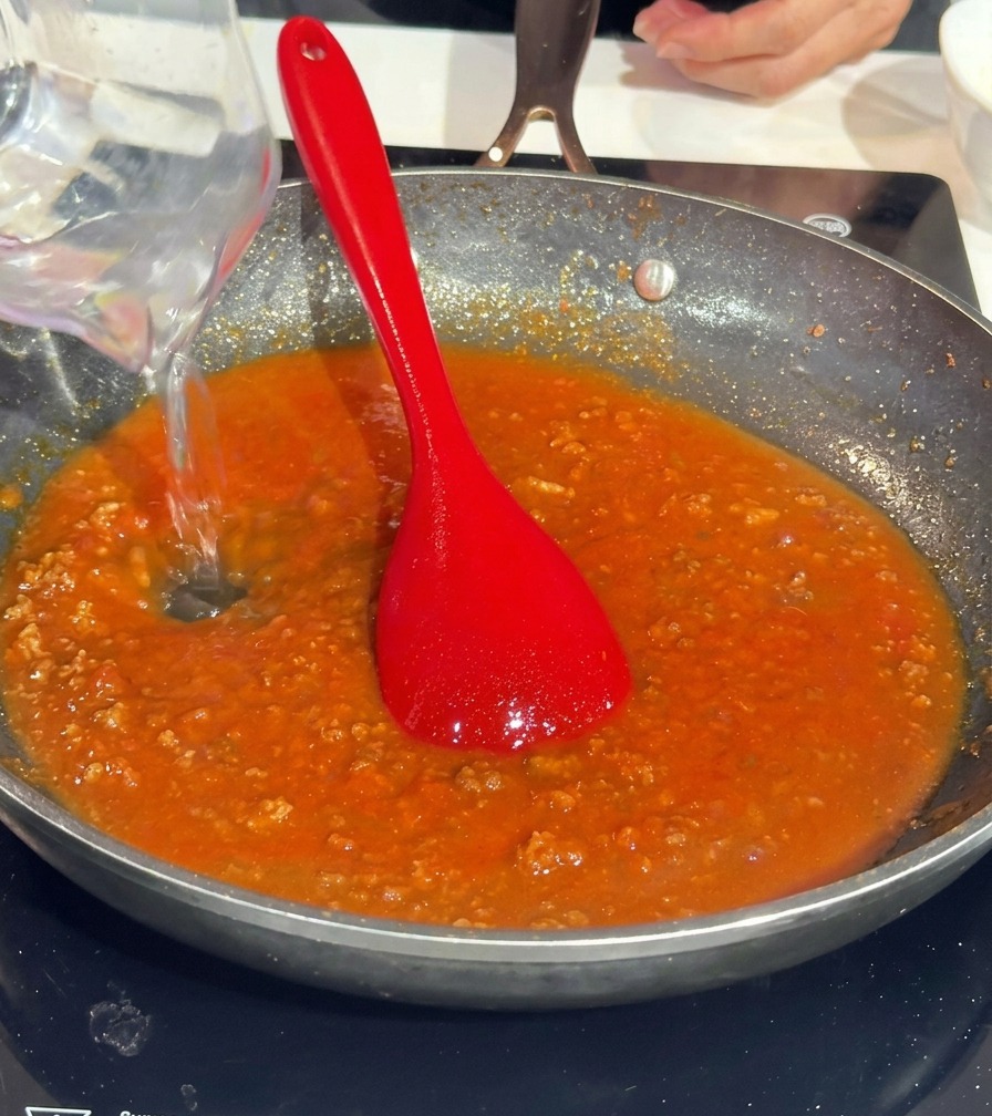 Verser de leau dun verre doseur transparent dans une poêle de sauce tomate à la viande.