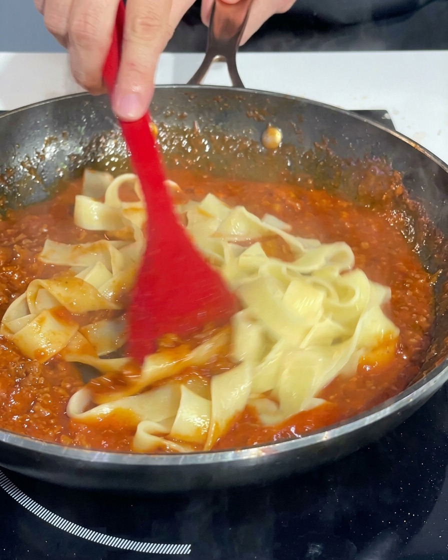 Une spatule rouge mélangeant les rubans de pâtes dans une poêle pour les enrober de sauce tomate à la viande.