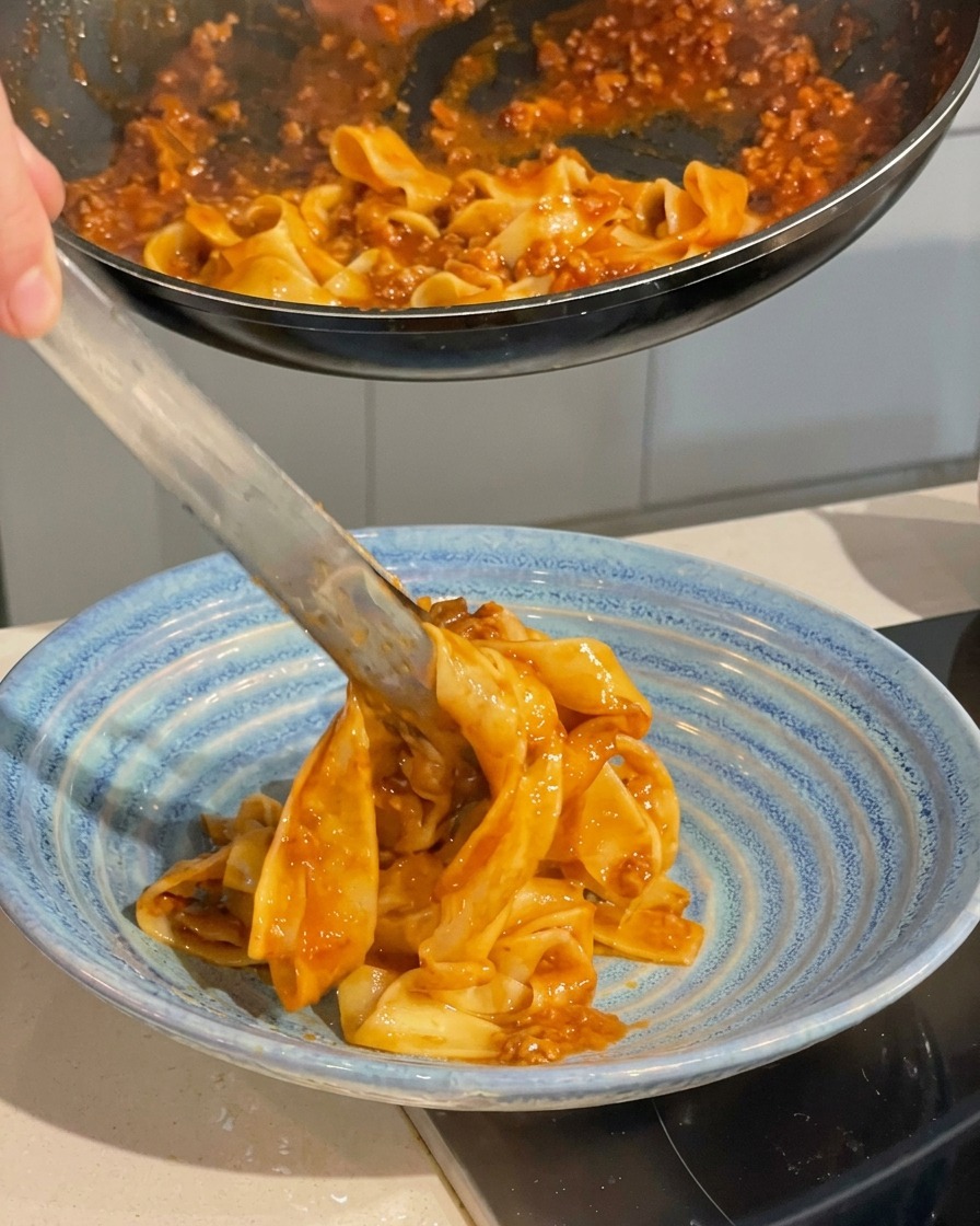 Une pince de cuisine soulevant des pâtes en sauce dune poêle pour les déposer dans un bol de service bleu profond.