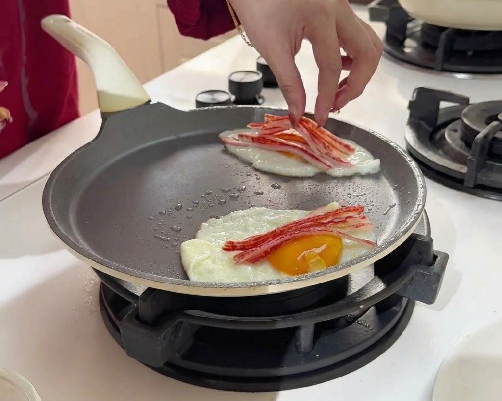 Placement de fines lamelles de surimi rouge et blanc sur la surface de deux œufs au plat en train de cuire.