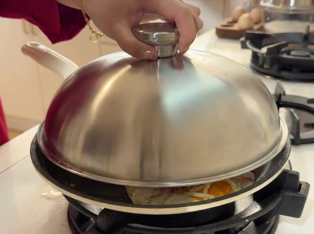 Une main posant un couvercle bombé en acier inoxydable sur une poêle contenant les œufs, le fromage et le surimi.