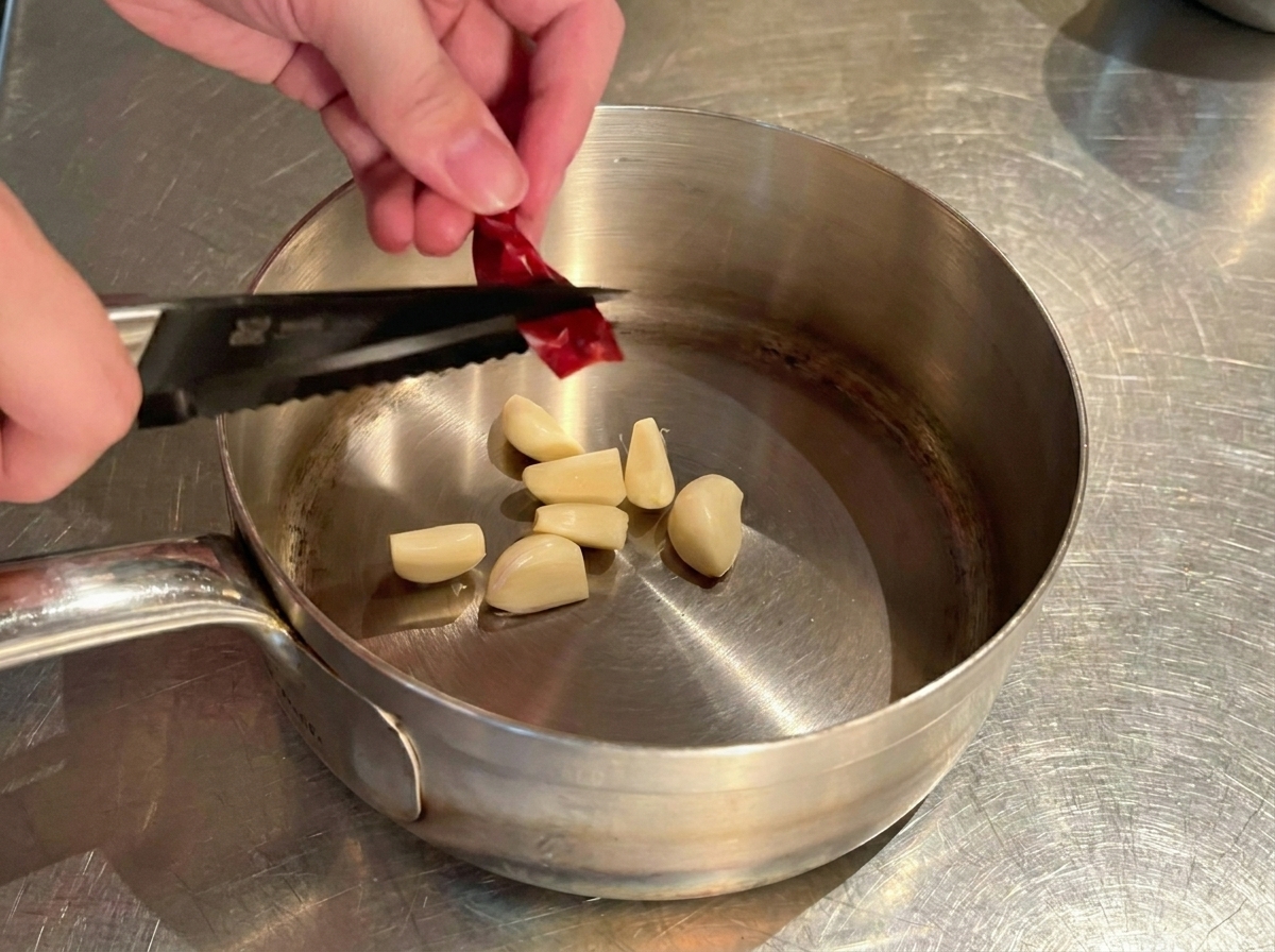 Mains découpant un piment rouge séché en petits morceaux au-dessus de lail dans la casserole.