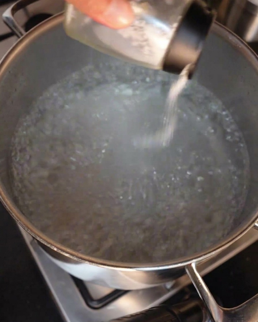 Une main saupoudrant du sel depuis une salière en verre dans une grande marmite en inox remplie deau à forte ébullition.