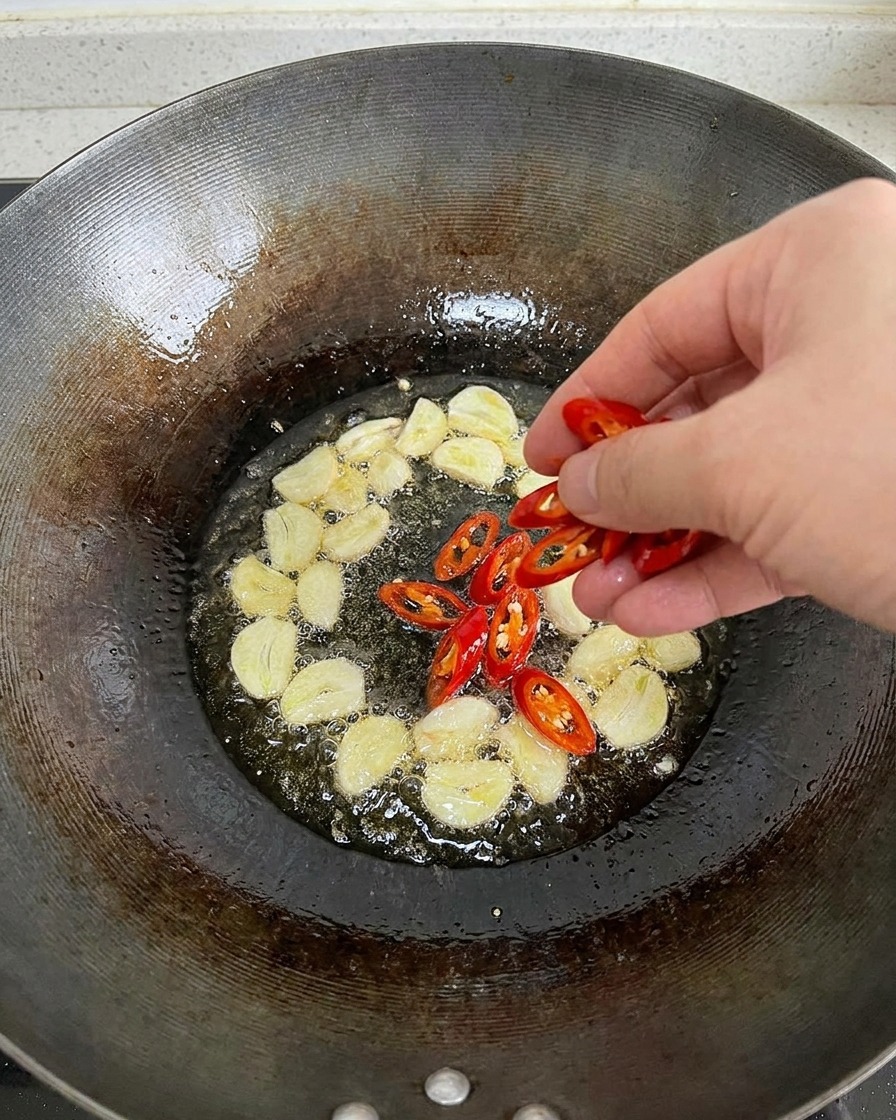 Une main jetant des piments rouges fraîchement émincés dans un wok dhuile chaude qui frémit déjà avec des tranches dail.