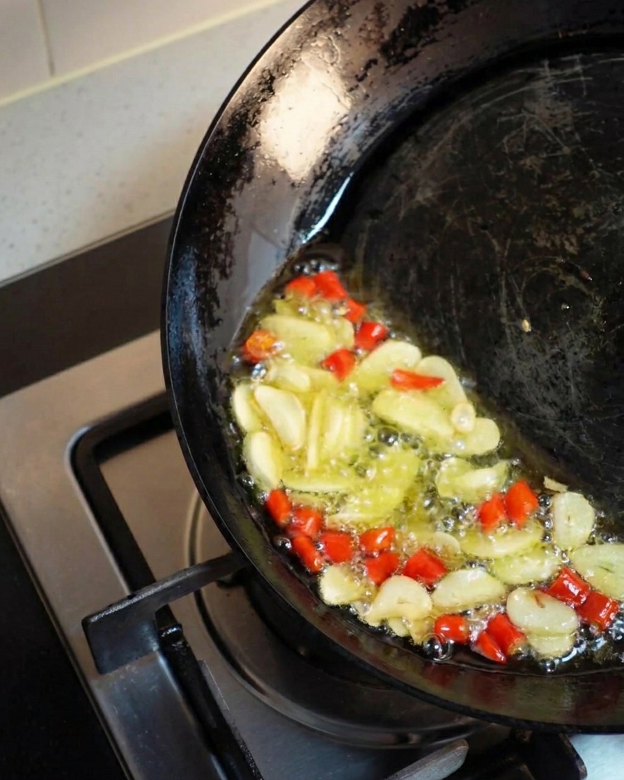 Un wok sombre incliné dun côté, rassemblant lhuile chaude et dorée où des tranches dail et des piments rouges hachés frémissent et frisent vigoureusement.