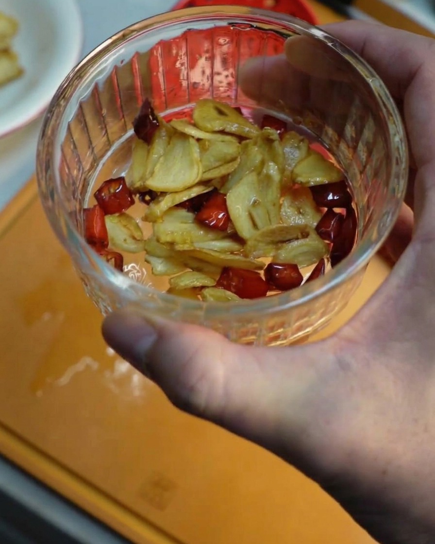 Une main tenant un petit bol en verre rempli de tranches dail frites croustillantes et de morceaux de piment rouge.