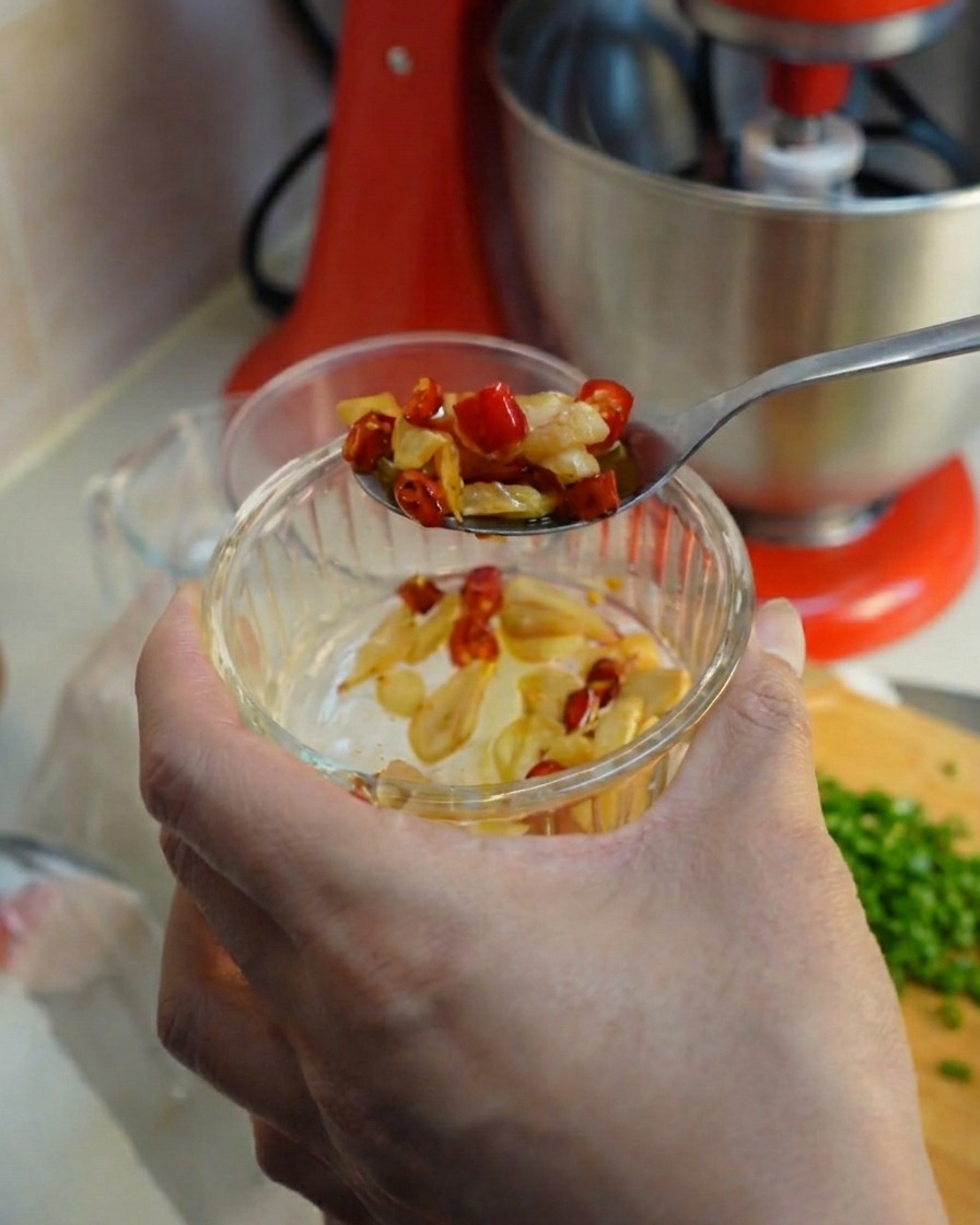 Une cuillère ramassant de lail frit et des tranches de piment rouge dans un petit bol en verre.