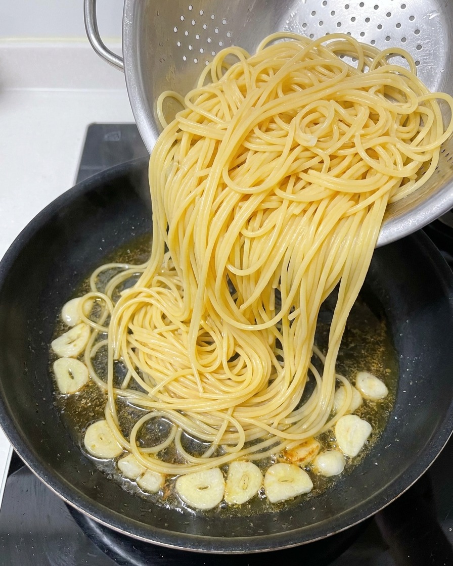 Des spaghettis cuits transférés depuis une passoire en métal vers une poêle contenant de lhuile infusée et des tranches dail frites.