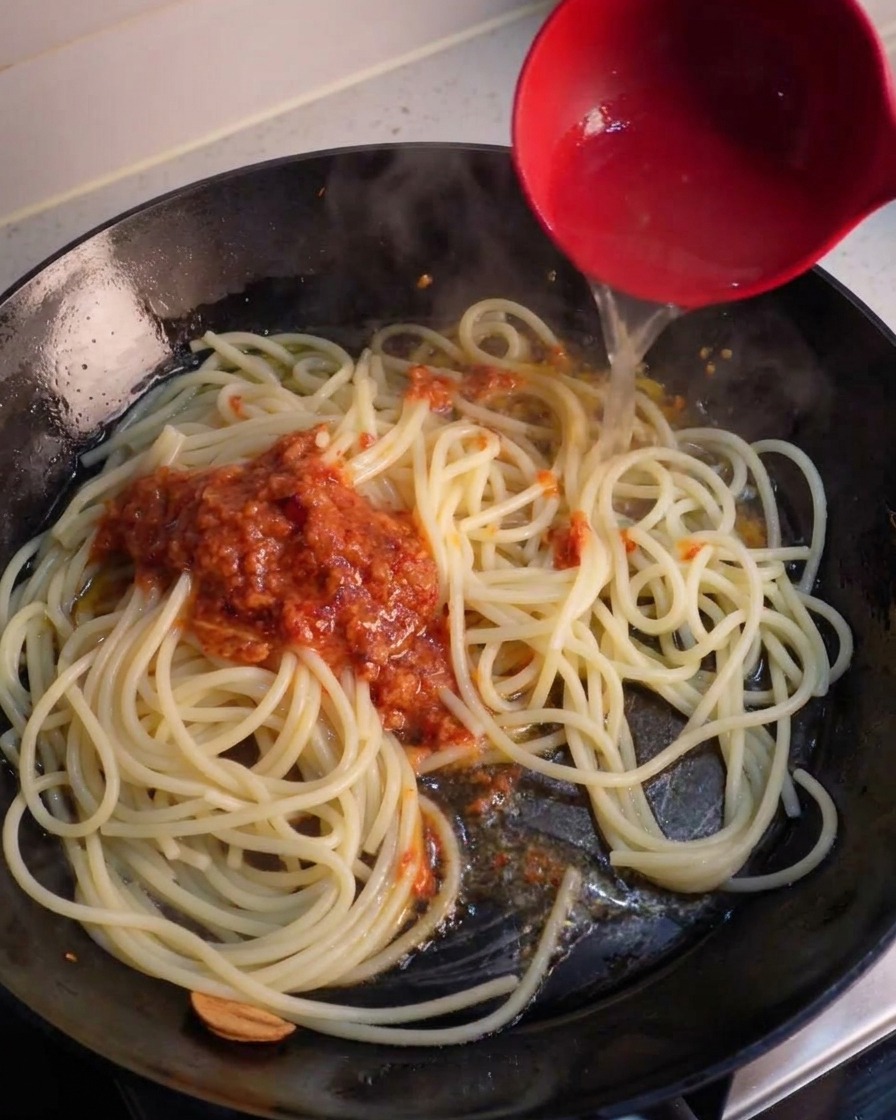 Une louche rouge versant leau claire de cuisson des pâtes dans une poêle sur des spaghettis et une cuillerée de sauce rouge mixée à lail et au piment.