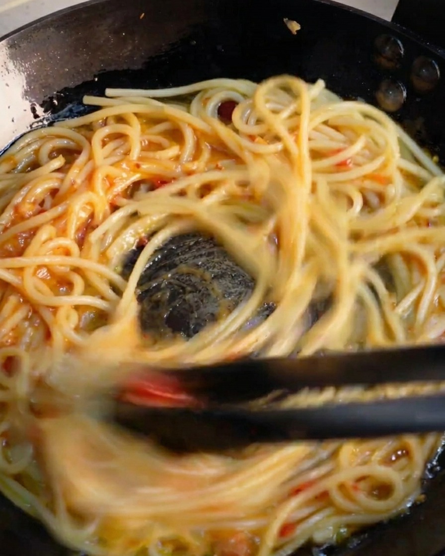 Des pinces noires mélangeant rapidement les spaghettis dans une poêle pour émulsionner la sauce vibrante à lail et au piment.