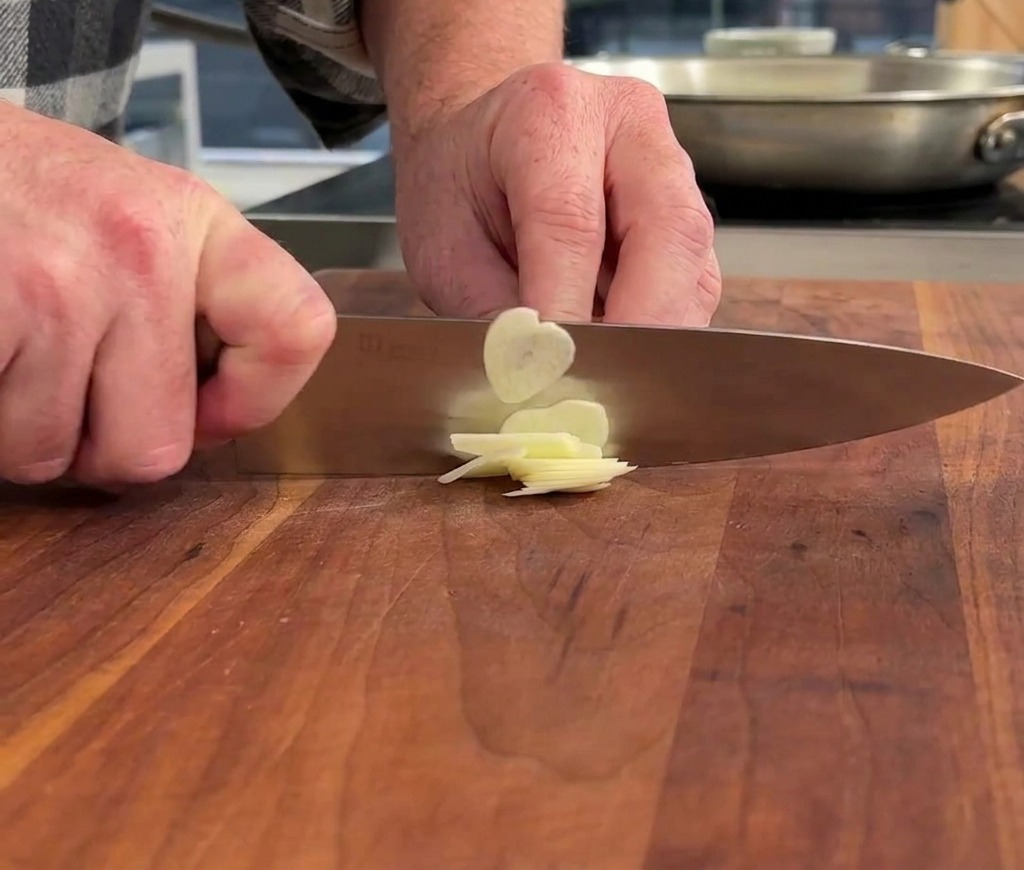 Mains utilisant un couteau de chef pour trancher finement une gousse dail sur une planche à découper en bois.