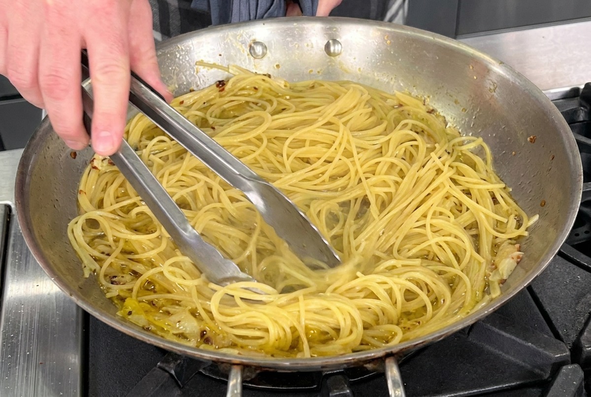 Utilisation de pinces pour mélanger les spaghetti dans une poêle en inox avec lhuile, lail et le piment.