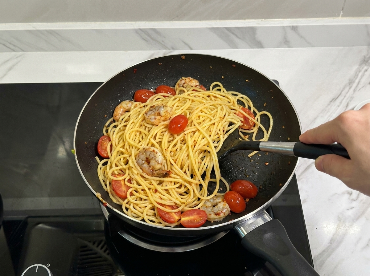 Une spatule noire mélangeant des spaghetti, des crevettes assaisonnées et des tomates cerises dans une poêle.