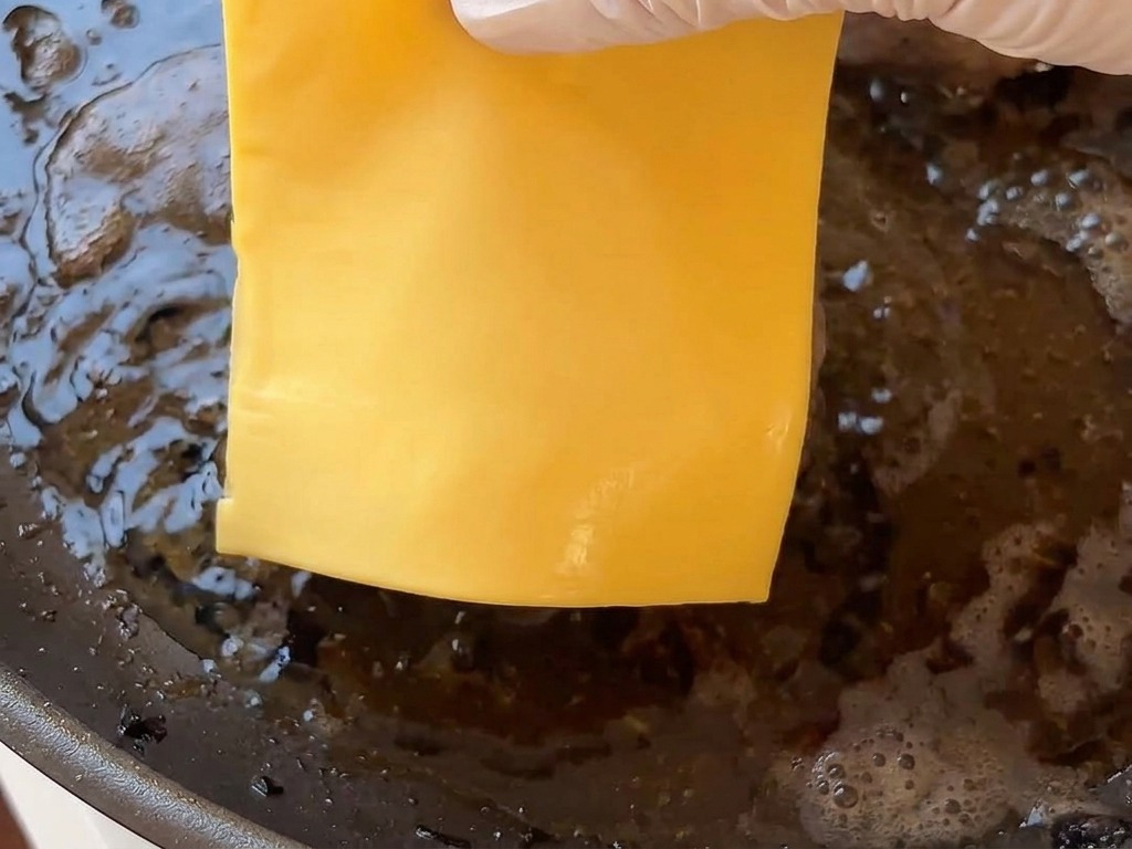 Une main gantée déposant une tranche de fromage carrée sur un steak de burger chaud dans la poêle.