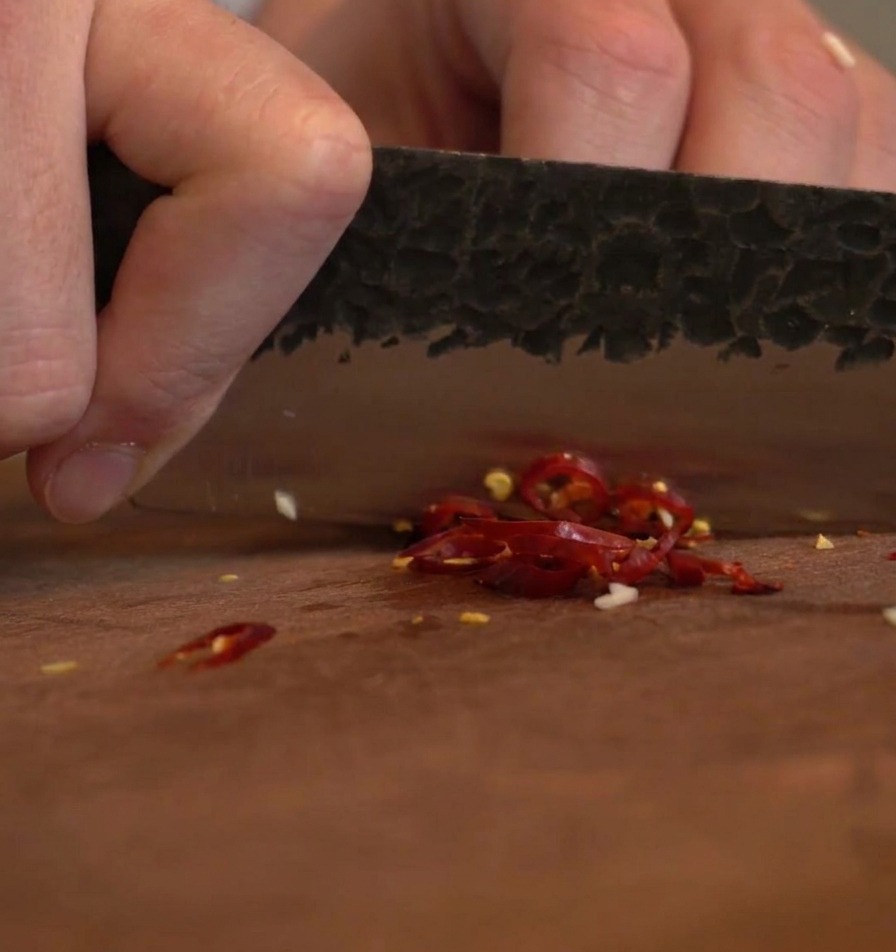 Une lame de couteau sombre hachant des piments rouges séchés sur une planche en bois.