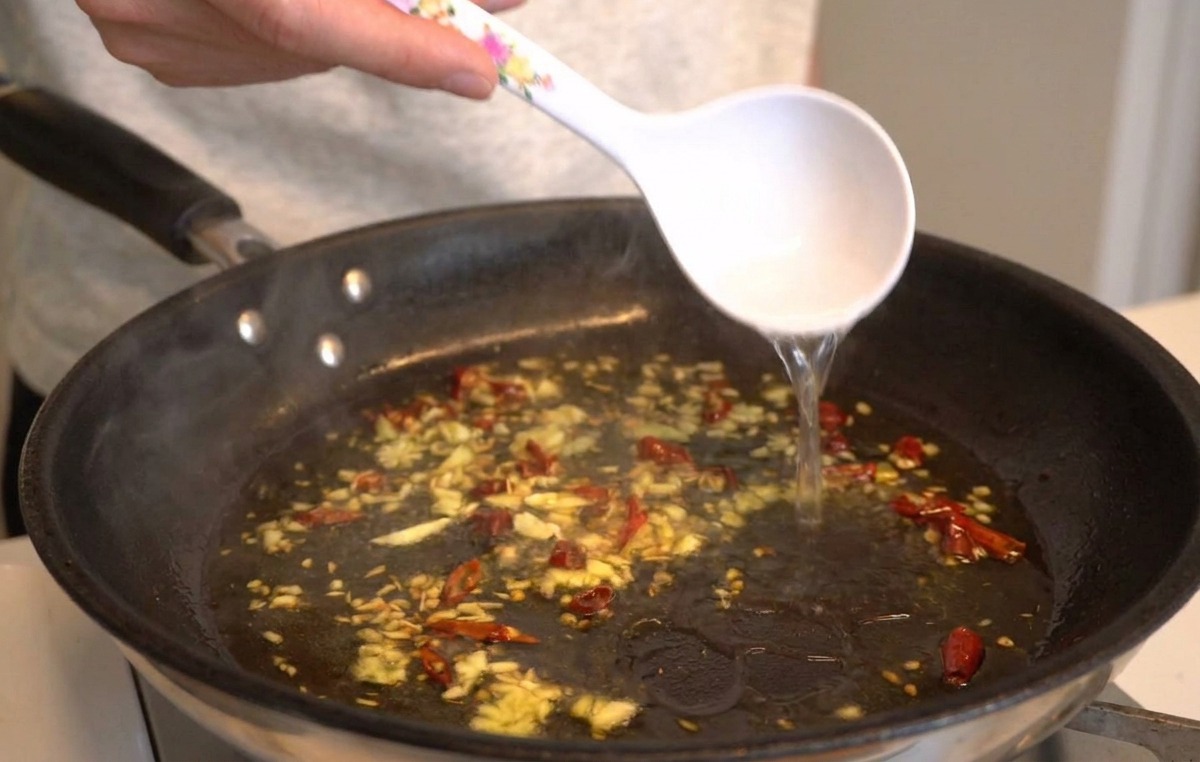 Une louche blanche versant de leau de cuisson amidonnée dans la poêle dhuile aillée.