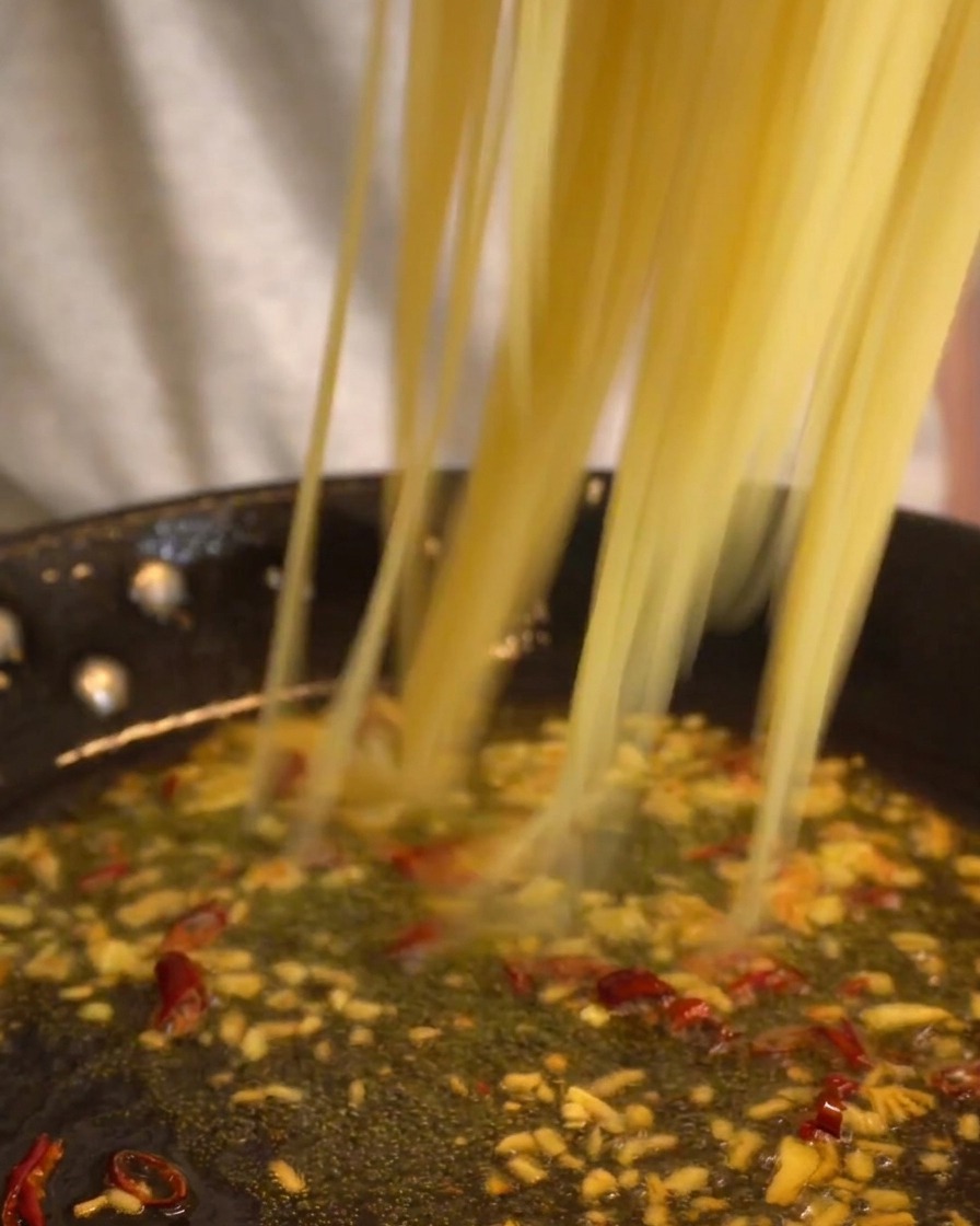 Longs brins de spaghetti déposés dans une poêle de sauce à lail et au piment frémissante.