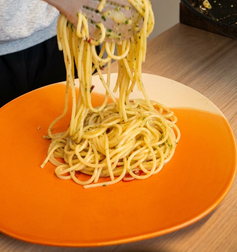 Pince soulevant et enroulant des spaghetti aglio e olio sur une assiette de service orange.