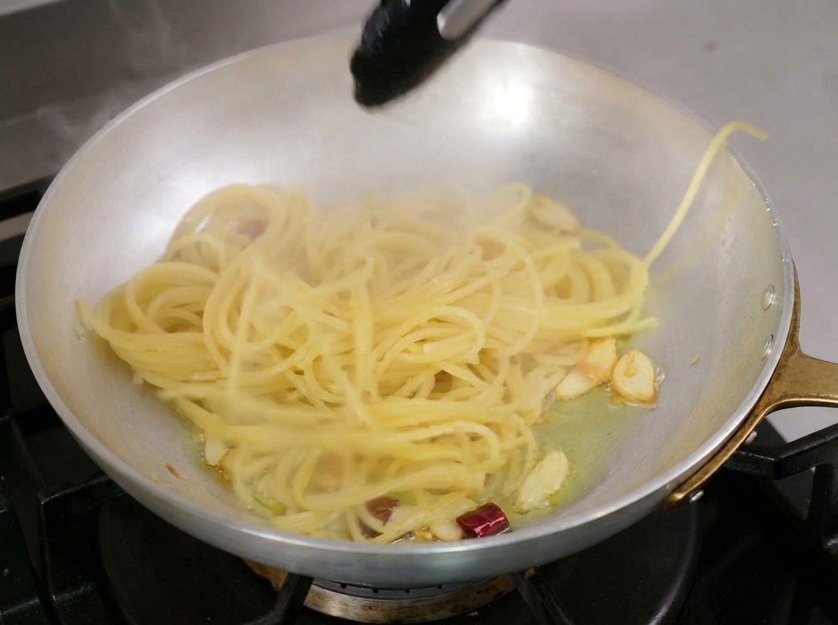 Pince de cuisine soulevant les spaghetti cuits pour les déposer dans la poêle dhuile parfumée.